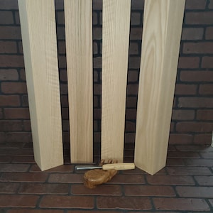 May include: Four unfinished wooden planks, likely oak, are stacked against a brick wall. A small wood carving of a hand holds a wood carving tool.