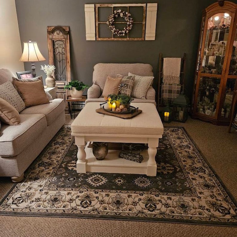 May include: A living room with a grey sofa, a white ottoman, and a patterned rug. The room is decorated with a wreath, a ladder, and a glass cabinet.
