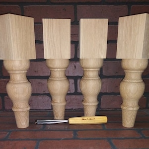 May include: Four unfinished wooden table legs with a turned, bulbous design. The legs are approximately 12 inches tall and are arranged in a row on a brick surface. A small wood carving tool is lying in front of the legs.