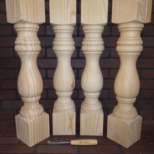 May include: Four unfinished wooden table legs with a turned design. The legs are square at the top and bottom and have a rounded, bulbous middle section. The legs are arranged in a row on a brick surface.