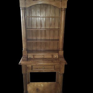 May include: A tall, wooden hutch with a top shelf and three drawers. The hutch is made of light brown wood and has a distressed finish. The hutch has a shelf on the bottom and a shelf on the top. The hutch has a total of four shelves.
