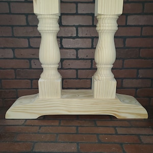 May include: A light-colored, unfinished wooden table base. The base features two ornate, turned columns connected by a rectangular base. The columns have a square top and bottom, with a rounded, carved design in the middle. The wood grain is visible.