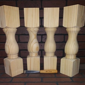 May include: Four unfinished wooden table legs with a turned design. The legs are square at the top and bottom and have a rounded, bulbous shape in the middle. The legs are made of light-coloured wood and are arranged in a row on a brick surface.