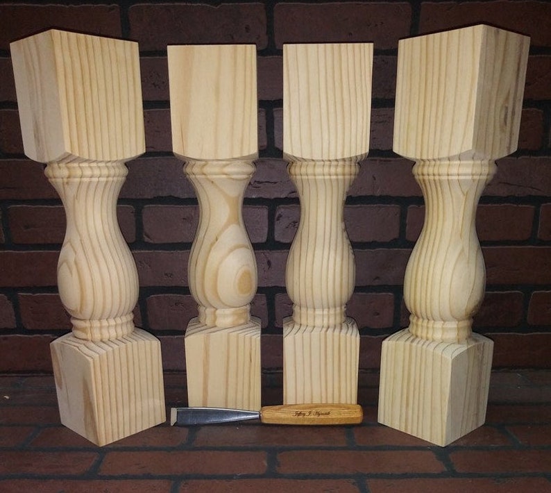 May include: Four unfinished wooden table legs with a turned design. The legs are square at the top and bottom and have a rounded, bulbous shape in the middle. The legs are made of light-coloured wood and are arranged in a row on a brick surface.