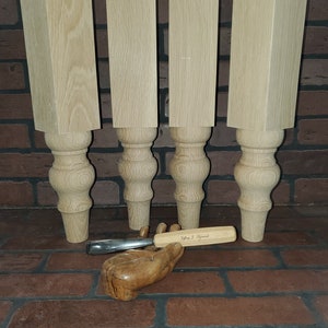 May include: Four unfinished wooden table legs with a turned design. The legs are approximately 10 inches tall and 3 inches wide. A wood carving tool is resting on a wooden hand.