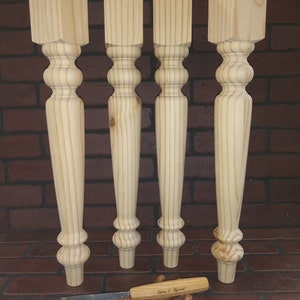 May include: Four unfinished wooden table legs with a turned design. The legs are square at the top and taper to a rounded bottom. The legs are arranged in a row on a brick surface.