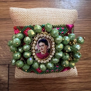 May include: A green beaded bracelet featuring a framed image of Frida Kahlo, surrounded by rhinestones. The bracelet is decorated with a colourful woven band.
