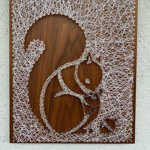 May include: A wooden string art piece featuring a squirrel design. The squirrel is crafted with silver-colored string against a dark brown wooden background. The background is filled with white string, creating a textured effect. The artwork is rectangular.