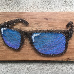 May include: A wooden wall art piece featuring a pair of sunglasses crafted from dark brown string and nails. The lenses are filled with blue and purple string, creating a vibrant, eye-catching design. The artwork is mounted on a rectangular, wood-grain background.