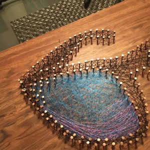 May include: A handmade string art piece in the shape of a stylized letter "G". The design features copper nails outlining the letter, with blue and purple threads woven within. The artwork is displayed on a wooden surface.