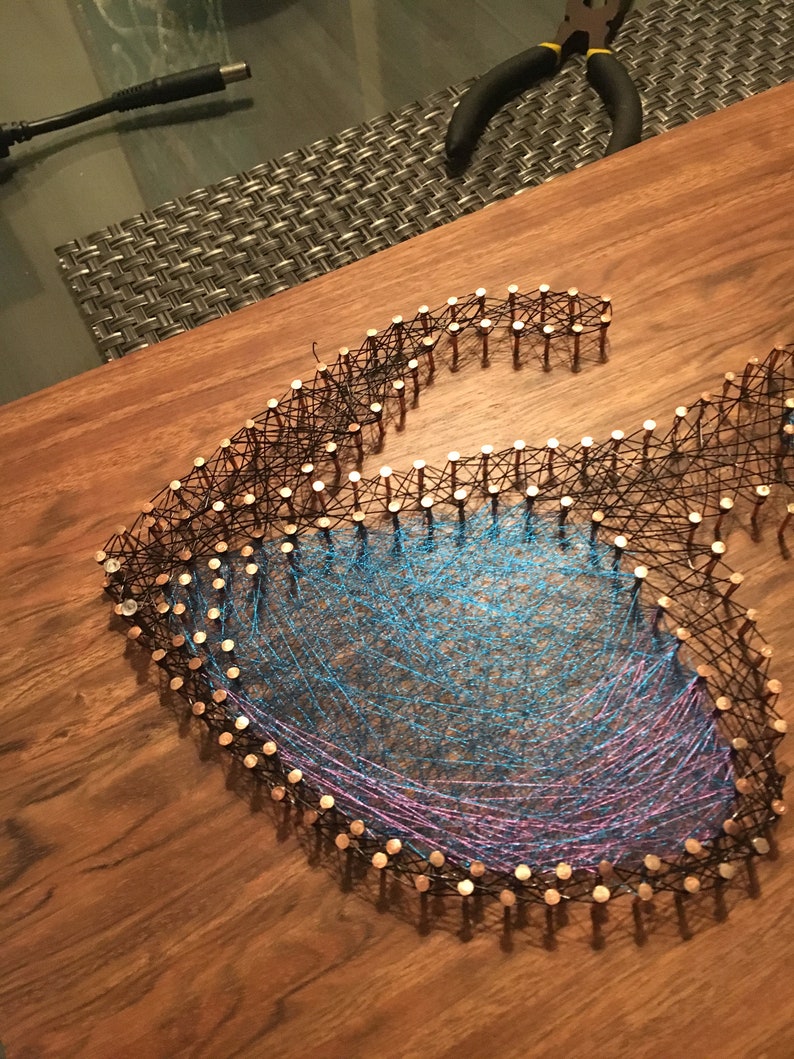 May include: A handmade string art piece in the shape of a stylized letter "G". The design features copper nails outlining the letter, with blue and purple threads woven within. The artwork is displayed on a wooden surface.