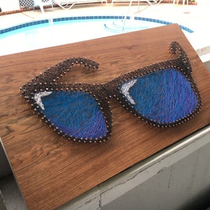 May include: A wooden art piece featuring a pair of sunglasses crafted with nails and blue thread. The sunglasses have blue lenses and are mounted on a brown wooden board. The artwork is displayed outdoors.
