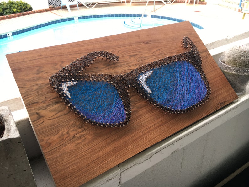 May include: A wooden art piece featuring a pair of sunglasses crafted with nails and blue thread. The sunglasses have blue lenses and are mounted on a brown wooden board. The artwork is displayed outdoors.