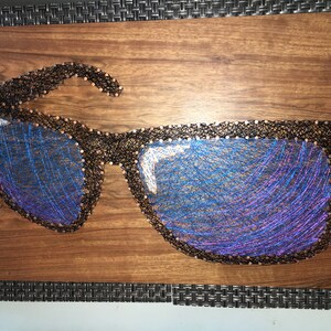 May include: A wooden art piece featuring a pair of sunglasses crafted with black thread and nails. The lenses are filled with blue and purple thread, creating a reflective effect. The artwork is mounted on a brown wooden board.
