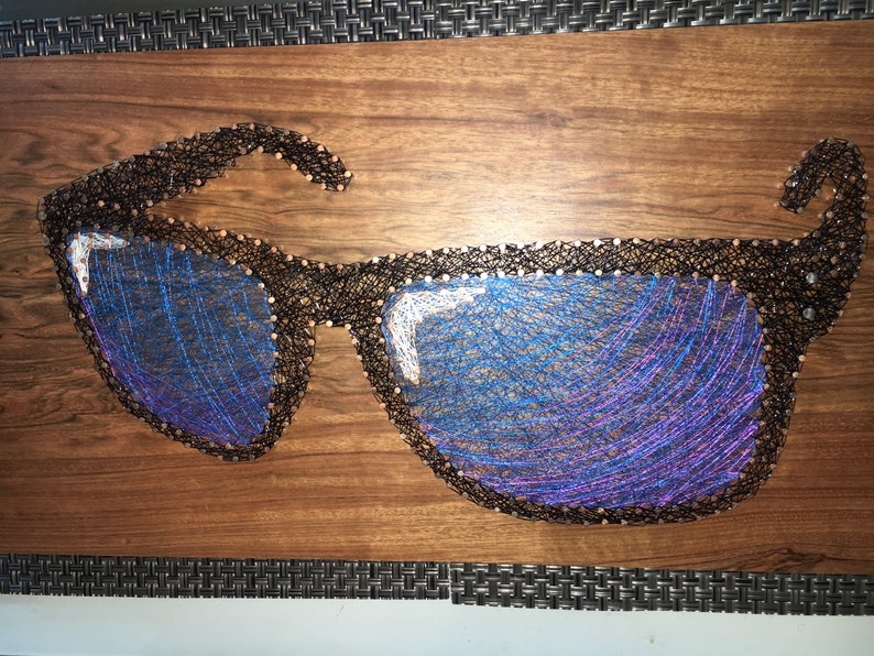 May include: A wooden art piece featuring a pair of sunglasses crafted with black thread and nails. The lenses are filled with blue and purple thread, creating a reflective effect. The artwork is mounted on a brown wooden board.