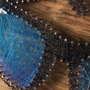 May include: A handmade string art piece depicting a pair of sunglasses. The sunglasses are crafted with black string outlining the frames and blue string filling the lenses. The piece is mounted on a wooden surface.