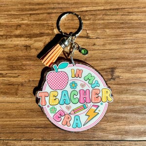 May include: A round keychain with the text "IN MY TEACHER ERA" in colorful bubble letters. The keychain features an apple, pencil, and lightning bolt graphic. It has a silver keyring, a tassel, and a green charm.