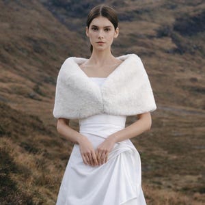 May include: A white faux fur stole draped over the shoulders, paired with a floor-length white satin gown featuring a straight neckline and a draped, gathered skirt detail that creates a soft, flowing silhouette.