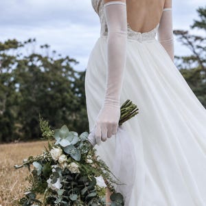 May include: A floor-length white wedding dress with a backless design and sheer elbow-length gloves. The garment is paired with a large bridal bouquet featuring white roses, green eucalyptus leaves, and ferns, tied with a light gray ribbon.