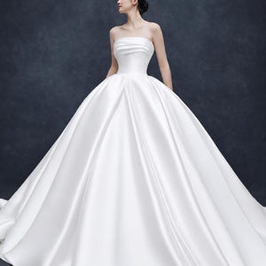 May include: A floor-length white strapless ball gown featuring a fitted bodice with horizontal pleating at the bust and a voluminous, full skirt with a smooth, satin-like finish that extends to the floor in a wide, circular silhouette.
