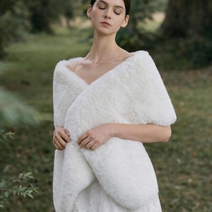 May include: A white faux fur wrap featuring a soft, plush texture and a wide, folded design that drapes across the shoulders and chest. The wrap is worn over a white lace garment, providing full coverage of the upper torso and arms.