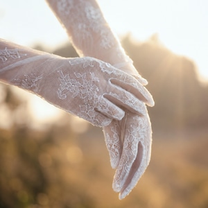 K&ouml;nnte beinhalten: Ein Paar wei&szlig;e Spitzenhandschuhe mit einem komplizierten Blumenmuster. Die Handschuhe reichen &uuml;ber das Handgelenk hinaus und bedecken die H&auml;nde und Finger mit einem zarten, halbtransparenten Netzstoff, der ein detailliertes florales Design aufweist.