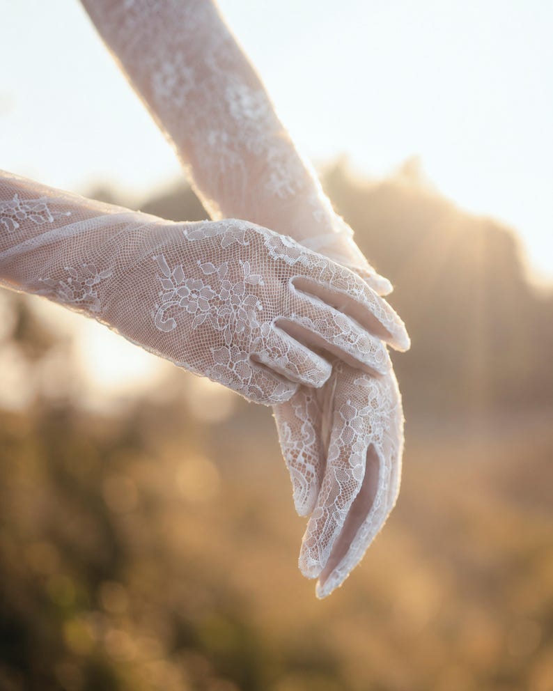 K&ouml;nnte beinhalten: Ein Paar wei&szlig;e Spitzenhandschuhe mit einem komplizierten Blumenmuster. Die Handschuhe reichen &uuml;ber das Handgelenk hinaus und bedecken die H&auml;nde und Finger mit einem zarten, halbtransparenten Netzstoff, der ein detailliertes florales Design aufweist.