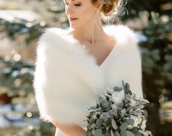 May include: A white faux fur stole worn over a white dress, featuring a soft, plush texture that drapes over the shoulders. The garment is paired with a bouquet of white flowers and green eucalyptus leaves, creating a clean, bright aesthetic.