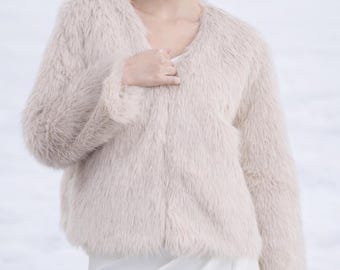 May include: A beige faux fur jacket with long sleeves and a collarless, open front design. The garment features a soft, shaggy texture throughout, providing a consistent light tan color. It is worn over a plain white garment with a smooth fabric finish.