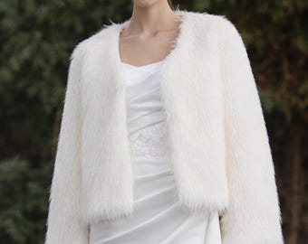 May include: A long-sleeved, open-front jacket made of white faux fur with a soft, shaggy texture. The jacket features a collarless, rounded neckline and a cropped length, worn over a smooth, white satin dress with subtle draped fabric details.