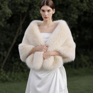 May include: A cream-colored faux fur stole draped over the shoulders, featuring a thick, plush texture and long, soft fibers. The stole is worn over a smooth, white satin dress with a strapless neckline and a draped, layered skirt design.