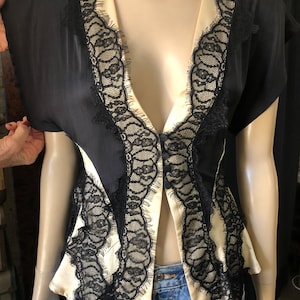 May include: Black and cream-colored blouse with a deep V-neckline. The blouse features black lace trim along the edges and a satin-like cream-colored panel down the center. The sleeves are short and the fabric appears to be silky.