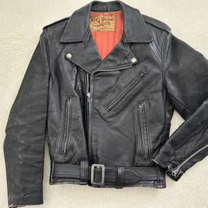 May include: Black leather motorcycle jacket with an asymmetrical zip, notched collar, and multiple zipped pockets. It has a belt at the waist and a red lining. The label reads "Kit Carson". The jacket is a classic biker style.