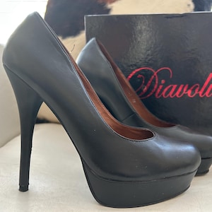 Könnte beinhalten: Ein Paar schwarze Plateau-High Heels mit Stiletto-Absätzen. Die Schuhe haben ein glänzendes Finish und eine runde Spitze. Eine schwarze Box mit dem Namen "Diavolina" in roter Schrift befindet sich im Hintergrund.