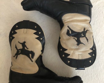 May include: A pair of black and cream-colored cowboy boots. The boots feature a black leather upper and a cream-colored foot with a black silhouette of a cowboy riding a horse. The boots have a pointed toe and a stacked heel.