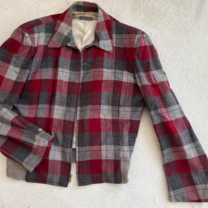 May include: A vintage jacket in a red, grey, and white plaid pattern. The jacket has a collared neckline, long sleeves, and a full zip closure. The lining is cream-coloured.