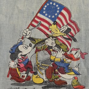 May include: A light blue fabric featuring a vintage illustration of Mickey Mouse, Goofy, and Donald Duck marching with a flag. The flag has red and white stripes and a blue field with stars. The text "Walt Disney Productions" is at the bottom.