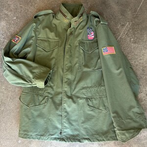 May include: An olive green military-style jacket with multiple pockets and patches. The jacket has a button-up front, a collar, and patches with various designs, including a peace sign and an American flag. The jacket is made of a durable fabric.