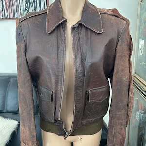 May include: A vintage brown leather jacket with a zipper closure, two front pockets, and a green ribbed hem and cuffs. The jacket has a classic collar and shoulder epaulets. The leather shows signs of wear, giving it a distressed look.