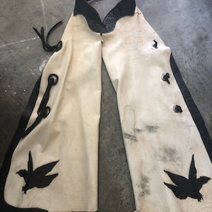 May include: Cream-colored leather chaps with black leather trim and decorative bird silhouettes. The chaps have a brown leather belt with a silver buckle and black leather accents. The chaps are designed to be worn over pants.