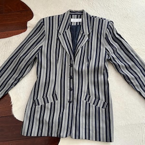 Puede incluir: Un blazer vintage a rayas verticales en azul marino y gris. El blazer tiene solapas, tres botones y dos bolsillos delanteros. La etiqueta dice "Jazelle".