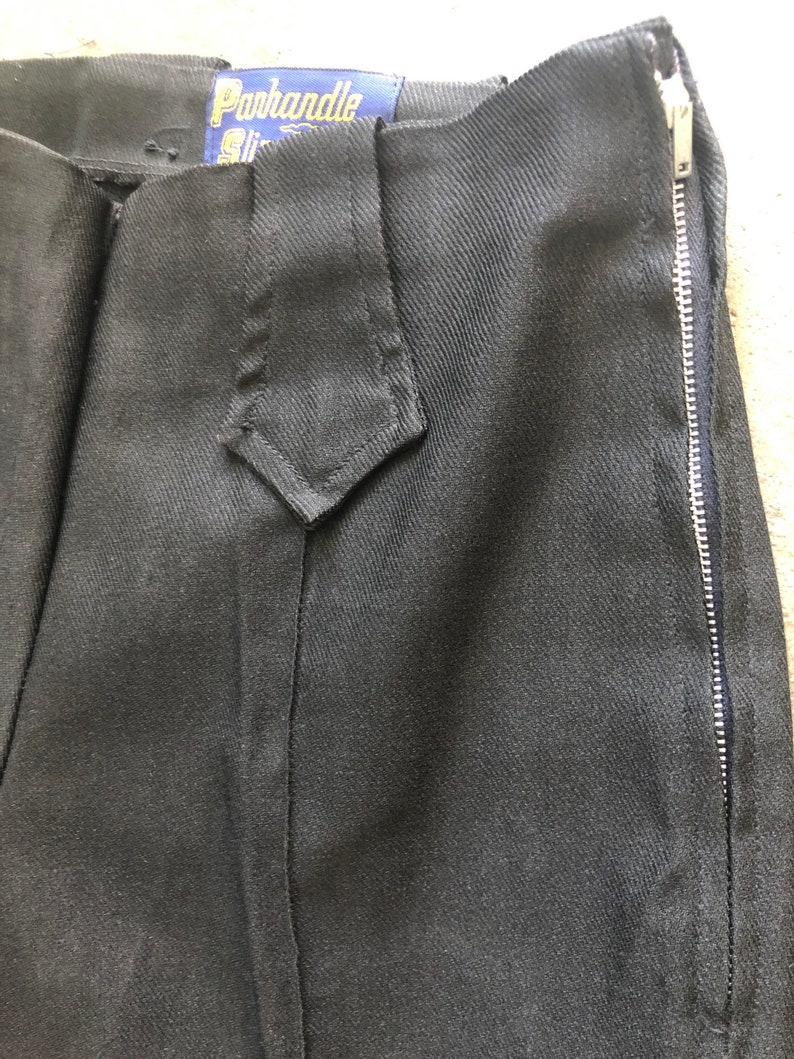 May include: Close-up of black pants with a side zipper. The pants have a diamond-shaped detail at the waist and a tag that reads "Parkhandle Slim". The fabric appears to be a sturdy, dark material.