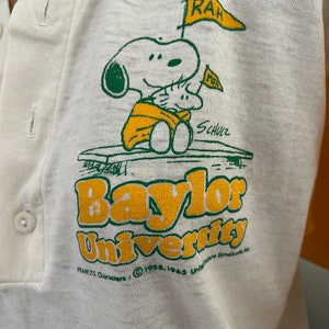 May include: Cream-colored polo shirt featuring a graphic of Snoopy holding flags with "RAH" on them. The shirt also displays "Baylor University" in yellow and green, with "Peanuts Characters" and copyright information.