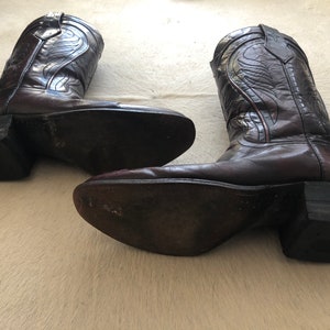 Puede incluir: Un par de botas de vaquero de cuero marr&oacute;n oscuro. Las botas tienen costuras decorativas y una puntera puntiaguda. Los tacones est&aacute;n apilados y las suelas muestran desgaste. Las botas se exhiben sobre una superficie clara.