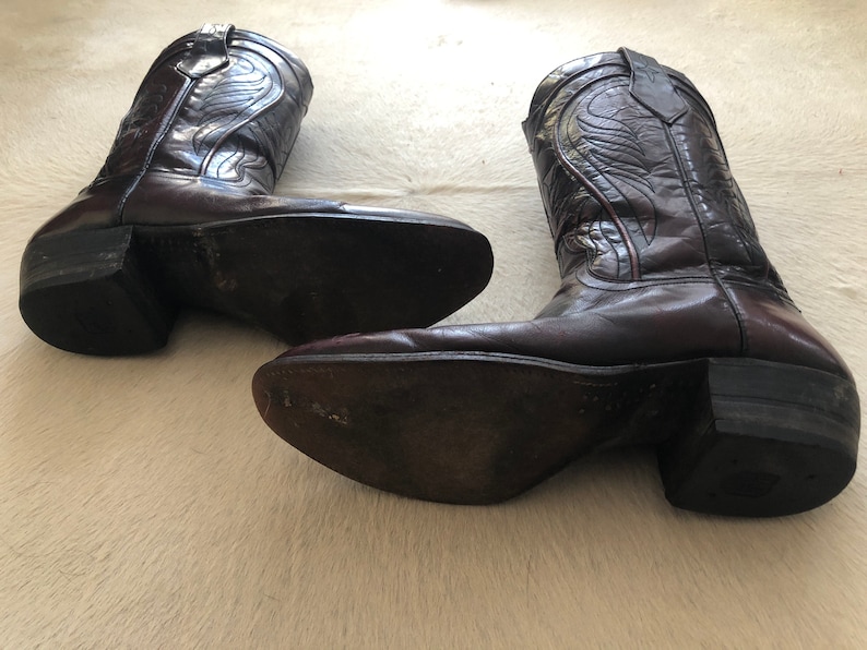 Puede incluir: Un par de botas de vaquero de cuero marr&oacute;n oscuro. Las botas tienen costuras decorativas y una puntera puntiaguda. Los tacones est&aacute;n apilados y las suelas muestran desgaste. Las botas se exhiben sobre una superficie clara.