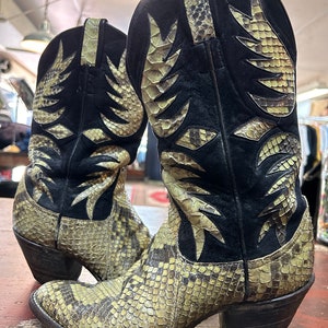 Puede incluir: Botas de vaquero con estampado de piel de serpiente y detalles en ante negro. Las botas tienen punta afilada y tacón grueso. El estampado de serpiente es una mezcla de colores beige, marrón y crema.