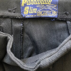 May include: Close-up of black denim pants with a blue label that reads "Panhandle Slim". The label also includes the text "Finest Quality Western Wear". The pants have a classic western fit.