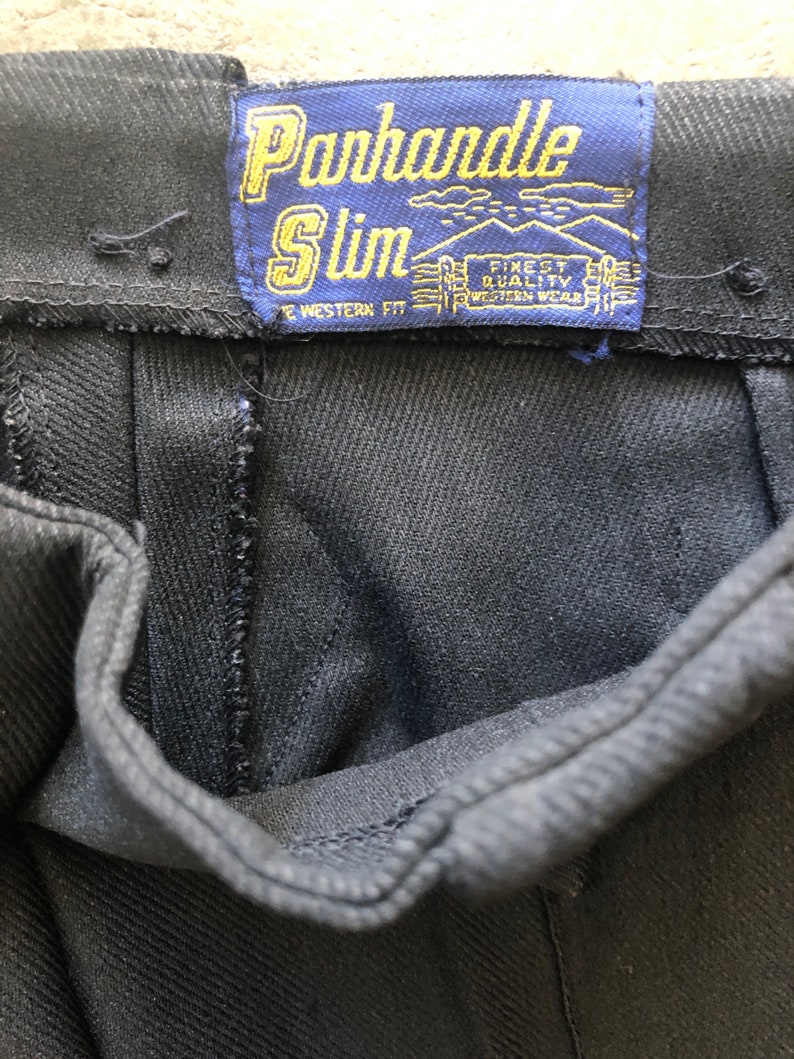 May include: Close-up of black denim pants with a blue label that reads "Panhandle Slim". The label also includes the text "Finest Quality Western Wear". The pants have a classic western fit.