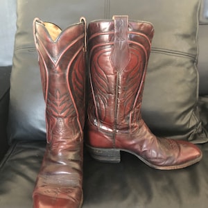 Puede incluir: Un par de botas de vaquero de cuero marr&oacute;n. Las botas tienen una punta afilada, costuras decorativas y una trabilla. El cuero tiene un acabado brillante y un tono marr&oacute;n rojizo. Las botas est&aacute;n sobre una superficie negra.