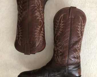 Può includere: Un paio di stivali da cowboy in pelle marrone. Gli stivali presentano cuciture decorative e una punta affusolata. La pelle ha un aspetto invecchiato. Gli stivali sono un classico stile Western.